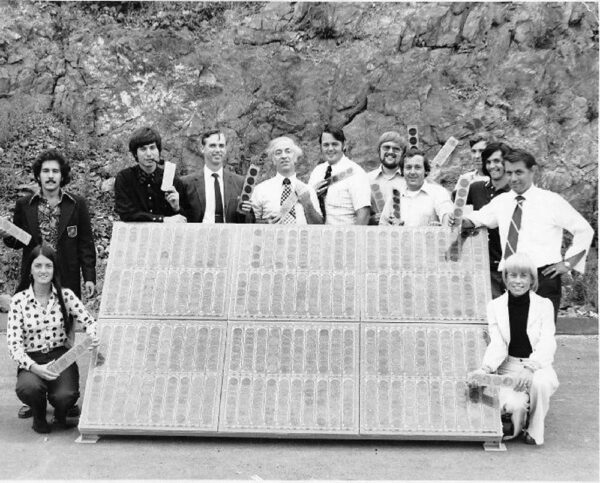 A group of people stands behind a small array of early solar modules mounted on the ground.