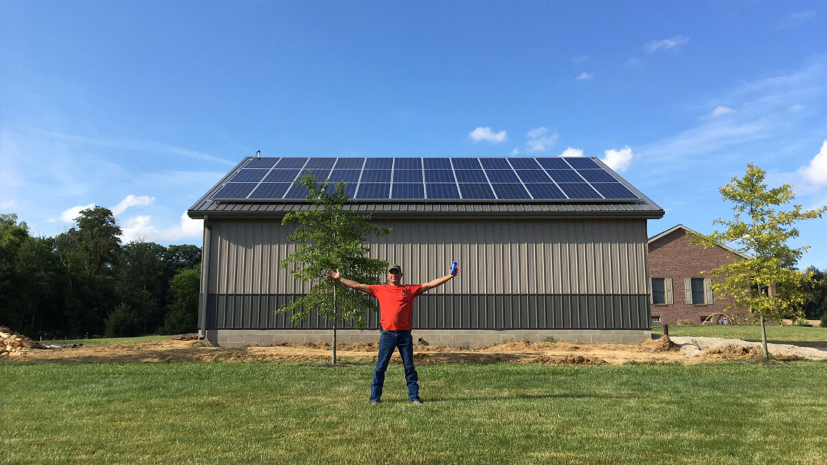 A man standing with arms outstretched in front of a large outbuilding with a completed solar installation on it.